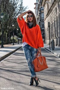 Модель, позирующая на улице, в джинсах с дырками, оранжевой сумкой и свитере