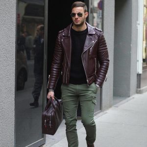 Мужчина, идущий по улице в бордовой куртке косухе, в зеленых брюках и с сумкой в руке